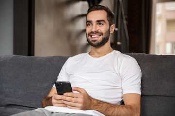 Photo of caucasian man holding and using smartphone while sitting on sofa in living room