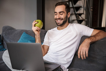 Photo of happy bachelor using silver laptop while sitting on couch in living room