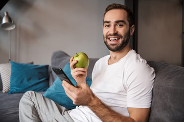 Photo of unshaved bachelor holding and using cell phone while sitting on couch in living room