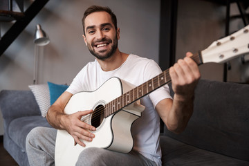 Photo of european man playing acoustic guitar while sitting on sofa in apartment