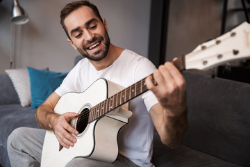 Photo of brunette man playing acoustic guitar while sitting on sofa in apartment