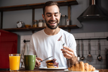 Image of brunette guy 30s eating food while having breakfast in modern apartment