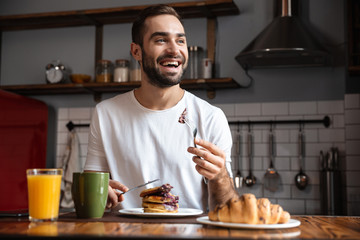 Image of caucasian guy 30s eating food while having breakfast in modern apartment