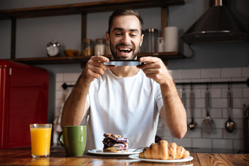 Portrait of happy man 30s holding smartphone and taking photo of food while having breakfast at home