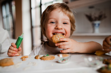 A small toddler boy sitting at the table, decorating and eating cakes at home.