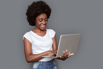 Fototapeta na wymiar Young woman standing isolated on gray with laptop browsing internet cheerful