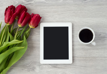 White tablet with a cup of coffee and red flowers lies on a white wooden table.