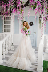 Fototapeta premium Elegant girl in a long white dress standing on the stairs with a bouquet in her hands
