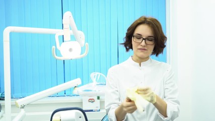 Young woman doctor wearing glasses and a white coat, putting on white medical glovesin dental clinic cabinet. A strict female dentist puts on gloves and looks into the camera, getting ready.