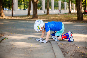 Little blonde boy 3 years old in white sport helmet and blue t-shirt riding on the roller-slates in the summer green park. Special problems with kid's eyes. Myopie, astigmatism, cross-eyed.