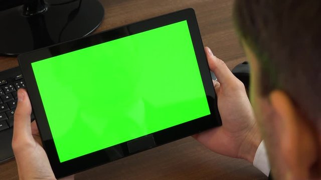 An office worker looks at a tablet with a green screen at a desk with a computer - closeup
