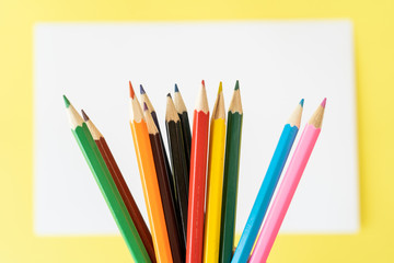 Close-up of a rainbow of colored pencils for child art above the white piece of paper on yellow background. Top view
