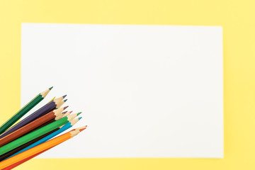 Bunch of colored pencils for child art above the white piece of paper in the center on yellow background
