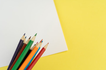 Bunch of colored pencils for child art above the white piece of paper on yellow background