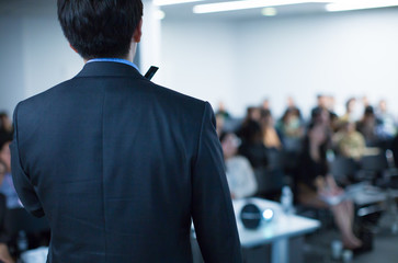 "Speaker on Stage at Conference Meeting Event. Presenter at Business Seminar Photo. Audience Watching a Manager Presentation. Blurred Image of Lecturer Presenting To Audience During Speech. "