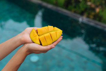 Close up slice of mango in female hands on background of blue swimming pool.