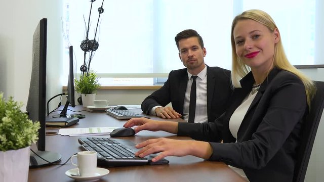 Two office workers, man and woman, work on computers and nod at the camera with a smile