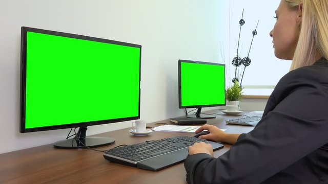 An office worker sits at a desk and works on a computer with a green screen - another desk with a green screen computer in the background