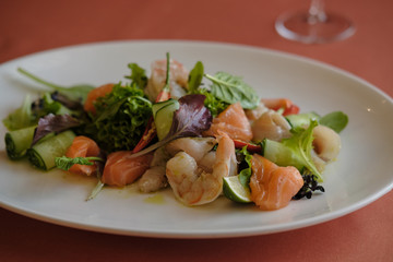Fresh delicatessen salmon salad (trout) with lettuce, basil, pepper, prawn and cucumber with mustard dressing and olive oil in a white plate. Selective focus
