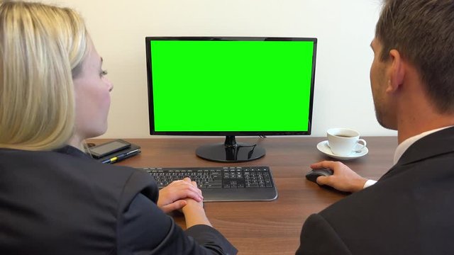 Two office workers, man and woman, sit at a desk and talk, the man works on a computer with a green screen - closeup