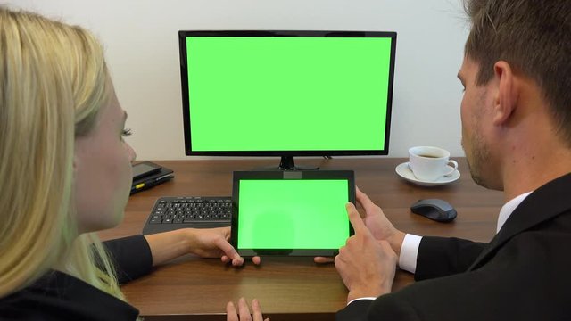 Two office workers, man and woman, sit at a desk and look at a computer and a tablet with green screens - closeup