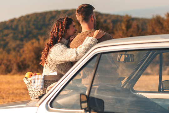 Beautiful young couple enjoying picnic time on the sunset. They are sitting on old fashioned car and looking at distance.