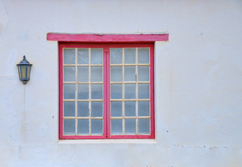 Minimalist capture of a single rural window on a white textured wall.  Window has small panels, and a red window frame. There is a house light