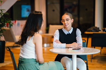 Two magnificent ladies discussing a record inside a bistro. They are talking about certain exercises while sitting on a table.