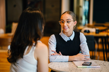 Two magnificent ladies discussing a record inside a bistro. They are talking about certain exercises while sitting on a table.
