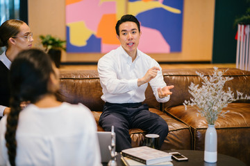 A handsome and confident Chinese Asian businessman sits on the chair and has a chat with his team during the day. He is enjoying a hot beverage while he talks animatedly to his colleagues.
