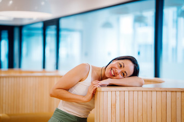 Young well dressed Eurasian woman smiling broadly in front view while resting her head on top of a modern styled room divider at a bright office. Image is taken in a mid-shot.