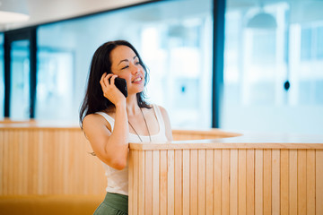 Portrait of a young,lovely Eurasian woman personnel having a chat and is smiling while holding her cellular phone. She is standing beside a stylish room divider.