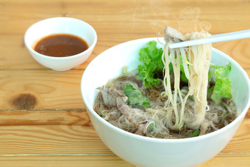 White noodle with Braised pork soup in white bowl with chopsticks and spoon and chili sauce, on brown wooden floor thai food
