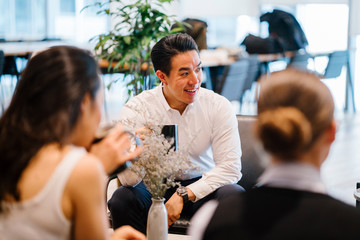 An attractive Chinese Asian man sits on the seat and stops for a moment to talk with his group amid the day. He is getting a charge out of a hot drink while he talks animatedly to his partners.