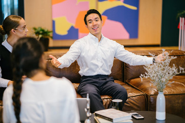 A youthful Asian Chinese expert is giving an introduction before his partners in the workplace. He is wearing a white polo shirt and dark jeans.