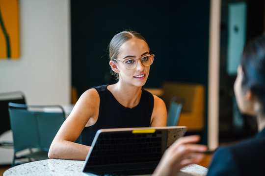 A young Caucasian woman discussing work with a team mate seriously inside a cafeteria. She is wearing eyeglasses and an elegant black dress. 