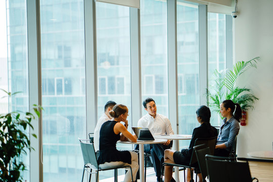 An assorted group lounge around a table in a gathering space to have a conference to talk about plans. The gathering is global with Asian and white colleagues and they are expertly dressed.