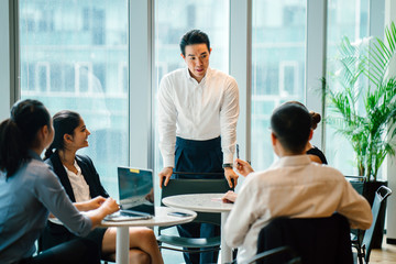 A dazzling Asian Chinese man situates a social occasion with his gathering in the midst of the day in the work environment. He is expertly wearing a shirt and pants and is motioning as he talks.