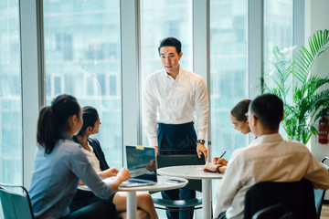 A gorgeous, sure, and fit Asian Chinese man seats a gathering with his group amid the day in the workplace. He is expertly wearing a shirt and pants and is motioning as he talks.