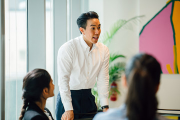 Obraz premium A good looking, confident and fit Asian Chinese man is presenting in a meeting with his team during the day in the office. He is professionally dressed in a polo and pants and is smiling as he speaks.