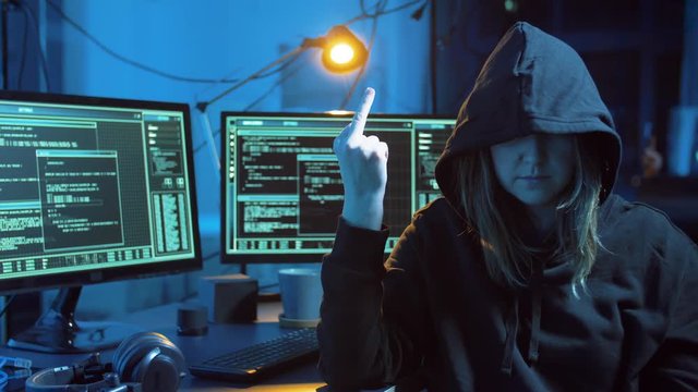 cybercrime, hacking and technology concept - female hacker committing cyber attack and showing middle finger next to computers in dark room