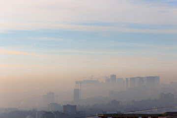 Construction cranes on new residential areas. Building new city on the on the edge of a high hill.Sunny foggy day. Houses silhouettes against blue sky