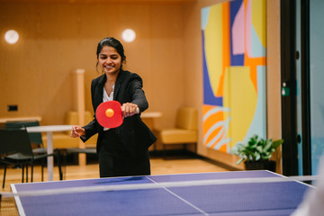 Portrait of a young and attractive Indian Asian woman in a suit playing table tennis with her colleague in the office during a break. She's having lots of fun. Image taken with a blurred art work. - obrazy, fototapety, plakaty