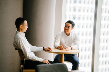 Portrait of two Asian Chinese businessmen chatting throughout the day in their office. They are professionally dressed in shirt and pants while sitting comfortably having a focused discussion.