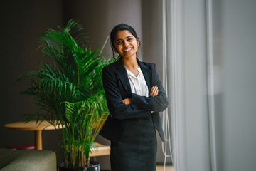 Image of a modern Asian Indian businesswoman standing by a glass window ledge in the office. She...