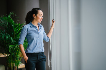 Fototapeta na wymiar Portrait of a beautiful young Asian woman looking at the view in her office from a glass window. She's clothed for the day in a casual blue shirt and black pants.
