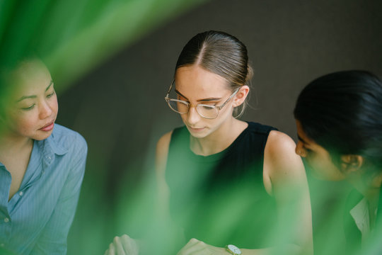 Three Professional Women Of Different Ethnic Backgrounds Sit Down And Have A Meeting Around A Table During The Day In A Conference Room. They Are All Dressed Professionally And Competently.