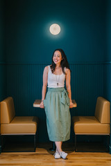 Portrait of an attractive and photogenic Asian woman entrepreneur leaning on a wooden table while smiling glamorously. She is wearing a casual and trendy white tank top and green skirt.