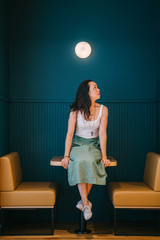 A beautiful and young Chinese Asian entrepreneur woman in a white tank top and green skirt. She is sitting on a table in her office place.