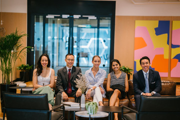 Group picture of an office-based business team sitting comfortably on the couch. All are professionally and competently dressed while smiling in front of the camera.